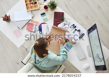 High angle view of young woman in hoodie sitting at desk with palettes and computer and drawing fashion clothing sketches in studio