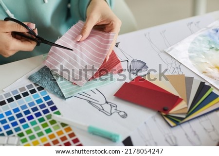 Close-up of unrecognizable woman sitting at desk with palettes and fashion sketches and cutting fabric in tailors studio