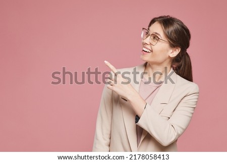 Smiling beautiful young female entrepreneur in fashionable jacket standing against pink background and pointing with finger at advertisement banner
