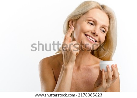 Skincare routine. Charming middle aged blonde 40s woman holding jar with anti-aging moisturizer, looking at the camera and applying cream isolated on white background, copy space. Advertising concept