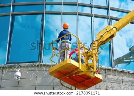 Female window cleaner cleaning glass windows on building at height in lifting platform. Worker polishing glass, window washing on office building in crane bucket. Window cleaners exterior glass facade Royalty-Free Stock Photo #2178091171