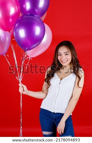 Cute and beautiful long dark hair Asian girl holding and playing with colorful air balloons with a funny and happy smile on red background, studio light shooting.