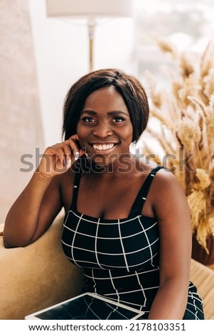 Happy stylish beautiful female student with hair looks at camera stands in cozy home interior. Smiling attractive young woman wears black dress headshot portrait.