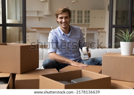 Portrait of smiling millennial man wrap cardboard packages using adhesive scotch, feel excited relocating to new house. Happy young Caucasian male renter pack boxes with belonging with tape dispenser. Royalty-Free Stock Photo #2178136639