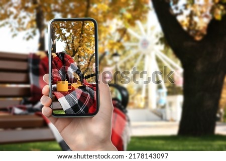 Woman taking picture of wooden bench with cup and plaid in park on autumn day, closeup