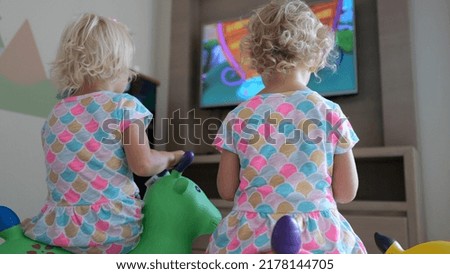 Children watch cartoons on TV in the playroom. Two blond girls sit on soft pillows in front of the TV and watch children's cartoons. The concept of teaching children TV from an early age. Royalty-Free Stock Photo #2178144705