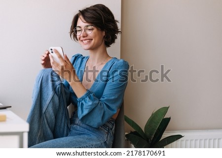 Charming woman holding phone at home . Caucasian smiling brunette woman looking and chatting on smartphone sitting on the floor. Concept of lifestyle, use technology  Royalty-Free Stock Photo #2178170691