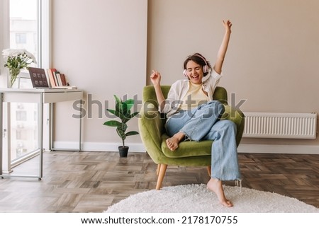 Young pretty woman listening music in headphones . Full view of funny caucasian woman having fun at home sitting on green chair. Concept of use technology  Royalty-Free Stock Photo #2178170755