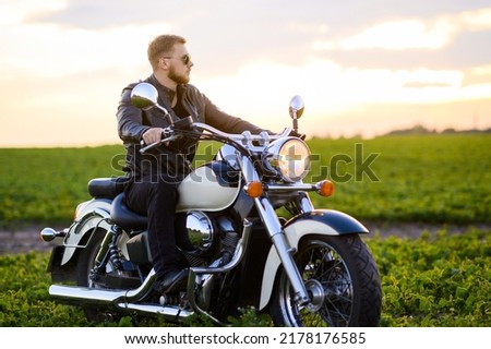 Young fair-haired brutal biker guy with a beard in sunglasses on a motorcycle in a field at sunset Royalty-Free Stock Photo #2178176585