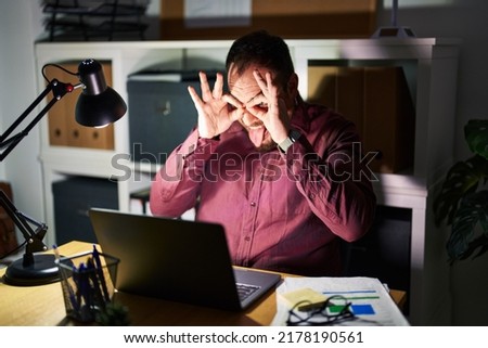 Plus size hispanic man with beard working at the office at night doing ok gesture like binoculars sticking tongue out, eyes looking through fingers. crazy expression. 