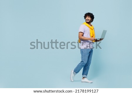 Full size body length side view young bearded Indian man 20s wears white t-shirt go move hold use work on laptop pc computer look aside isolated on plain pastel light blue background studio portrait