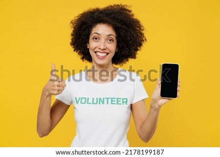 Young overjoyed fun woman of African American ethnicity wearing white volunteer t-shirt hold use mobile cell phone with blank screen workspace area show thumb up isolated on plain yellow background.