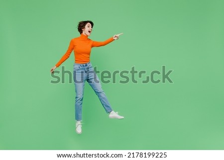 Full body young fun woman 20s wear orange turtleneck point index finger aside on workspace area mock up jump high isolated on plain pastel light green color background studio People lifestyle concept