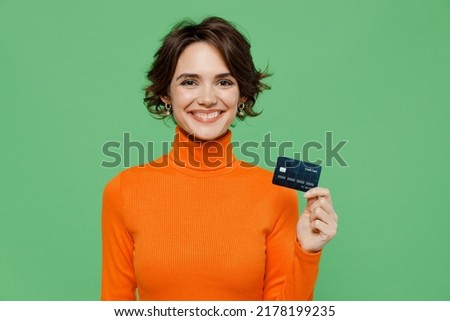 Young smiling happy cheerful fun woman 20s wear casual orange turtleneck hold in hand credit bank card isolated on plain pastel light green color background studio portrait. People lifestyle concept
