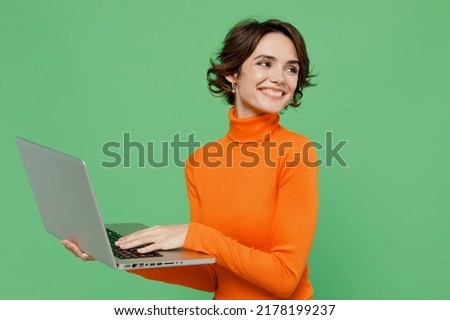 Young smiling copywriter happy fun woman 20s in orange turtleneck hold use work on laptop pc computer look aside on workspace area mock up isolated on plain pastel light green color background studio