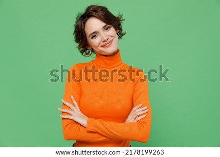 Young smiling happy confident woman 20s wear orange turtleneck hold hands crossed folded look camera isolated on plain pastel light green color background studio portrait. People lifestyle concept.