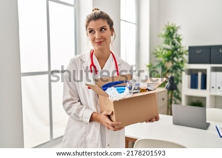 Young doctor woman holding box with medical items smiling looking to the side and staring away thinking. 