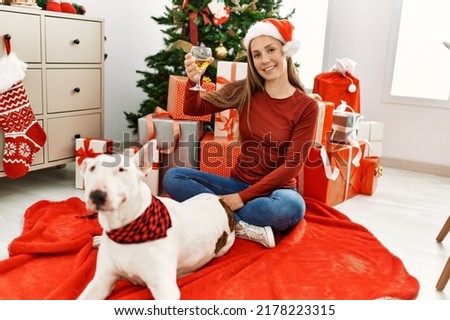 Young caucasian woman drinking champagne sitting with dog by christmas tree at home
