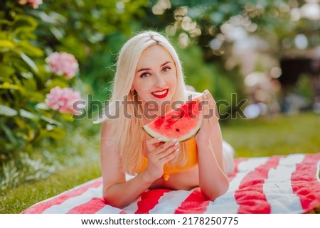 Pretty blond young woman in yellow bikini posing with slice of watermelon laying on red and white stripped towel in the garden and smiling.