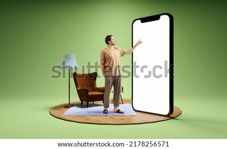 Pointing at device screen. Photo and 3d illustration of man standing next to huge 3d model of smartphone with empty white screen isolated on green background. Mockup for ad, text, design, logo Royalty-Free Stock Photo #2178256571