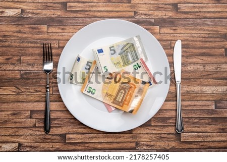 Euro banknotes lying on a dinner plate with cutlery on the table. Symbolic image for eating money notes. Food is getting more expensive due to inflation effects. Hunger caused by a crisis. Royalty-Free Stock Photo #2178257405