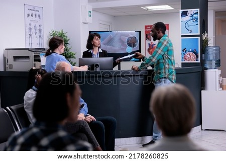 African american patient handing clipboard with completed form to hospital receptionist standing at front desk. Man attending doctor appointment in busy private clinic with diverse patients. Royalty-Free Stock Photo #2178260825