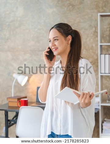 Smiling young Asian woman manager working remotely holding paper and reading financial documents Talking on a cell phone standing at a desk at home.