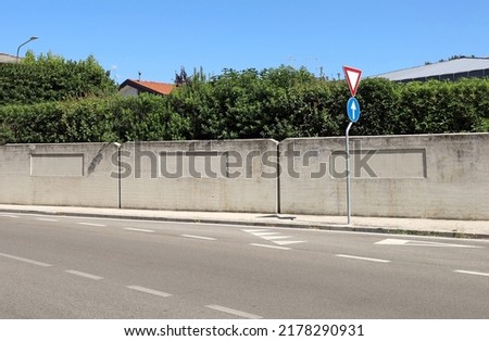 Give way and one way signs on a concrete sidewalk, Cement fence and hedge on behind, asphalt road with road markings in front. Background for copy space.