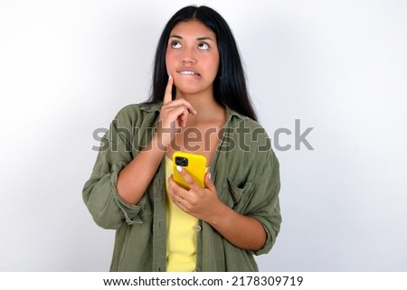 Image of a thinking dreaming Young hispanic woman wearing green jacket over white background using mobile phone and holding hand on face. Taking decisions and social media concept.