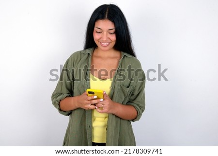Smiling Young hispanic woman wearing green jacket over white background using cell phone, messaging, being happy to text with friends, looking at smartphone. Modern technologies and communication.