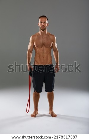 A topless sportsman performs fitness exercises with rubber band in a studio on gray background. Body-building. Athlete.