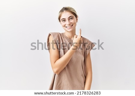 Young blonde woman standing over isolated background cheerful with a smile of face pointing with hand and finger up to the side with happy and natural expression on face 