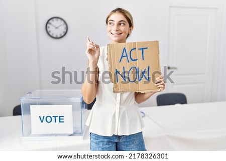 Young blonde woman at political election holding act now banner smiling happy pointing with hand and finger to the side 