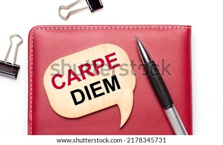 On a light background there are black paper clips, a pen, a burgundy notepad a wooden board with the text CARPE DIEM