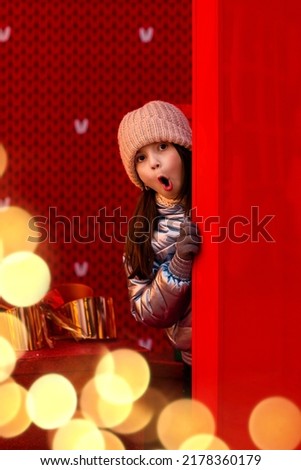 The girl peeks out from behind the red wall. New Year's location. Surprise on the girl's face. New Year Christmas. Winter. Holiday