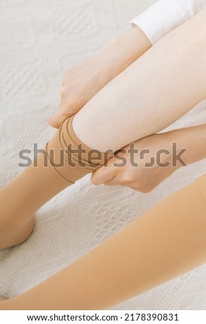 Beautiful woman putting on stocking indoors, closeup. Beautiful long female legs in stockings. Girl putting on stockings at home in a white room. Beige knee socks or socks.