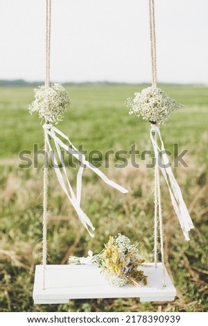 Wooden swing on a rope decorated with bouquets of wildflowers against the backdrop of a green field. Love story or wedding day concept. Romantic wallpaper banner or postcard. Royalty-Free Stock Photo #2178390939