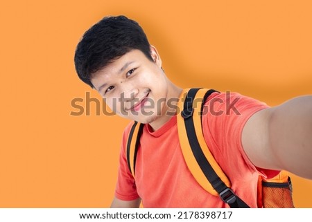 Portrait of an Asian man taking a selfie wearing a t-shirt with a backpack in studio orange tones. orange shade concept