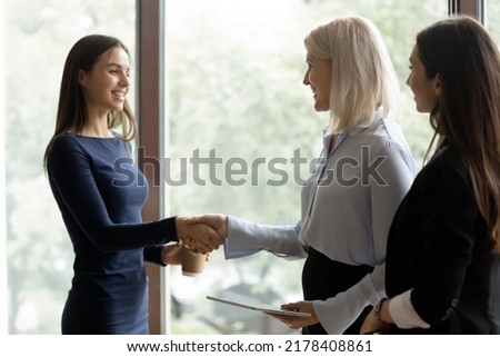 Young woman employee shaking hand of senior female colleague or business partner at work break in office getting acquainted, feeling grateful for help or assistance, congratulating with achievement Royalty-Free Stock Photo #2178408861
