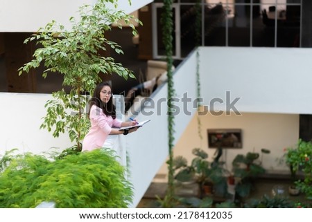 Beautiful business woman in glasses, in a pink suit, stands on a balcony surrounded by green plants with a notebook and a pen in her hands, looks at the camera