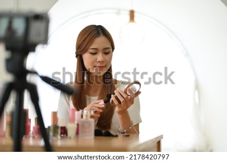 Pretty young Asian female freelance beauty blogger applying makeup, doing a makeup tutorial video, live-streaming her makeup content on social media.