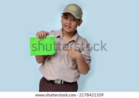 Asian boy in scouting uniform pointing at a green book, isolated white background