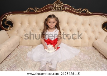 A little girl is waiting for the New Year and Christmas. A child in a white dress sits on a royal sofa with a cup of sweets in his hands. Celebration of the holiday. Wedding. Children's imagination. 