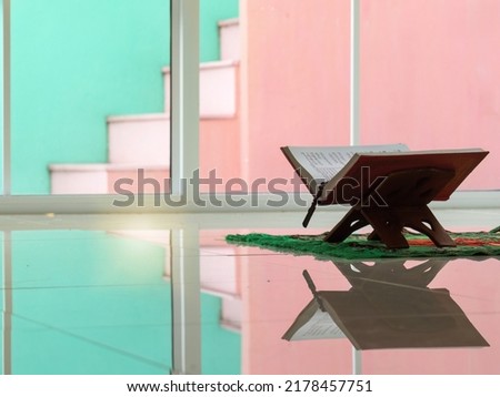 Rehal or wooden stand with open Quran on mat indoor with pink and green background.
