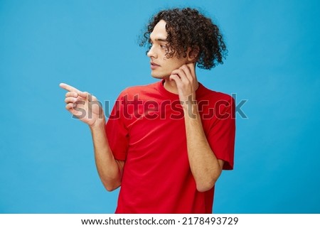 Excited shocked curly tanned Caucasian guy in basic red t-shirt point finger at free place looks aside posing isolated on over blue background. Lifestyle and Emotions concept. Good offer for ad