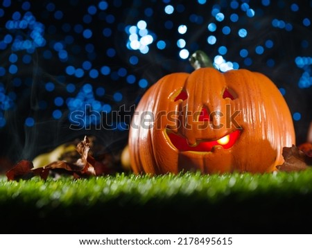 An orange pumpkin with a smile illuminated from within, fruits and cones on green grass against a blue starry sky. Halloween background. Advertising, banner, invitation.