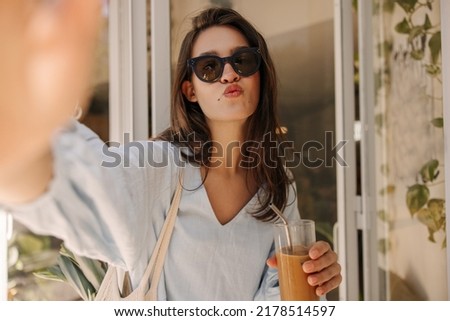 Cute young caucasian woman in sunglasses takes selfie, sends air kiss to camera. Brunette wear blue dress is holding glass of smoothie. Summer vacation concept.