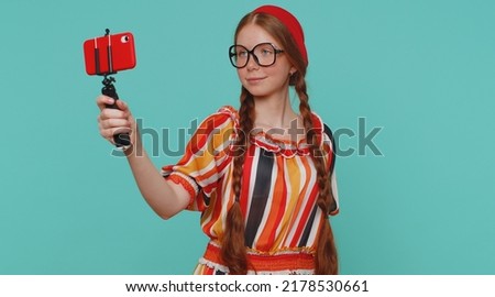 Redhead girl in dress traveler blogger taking selfie on mobile phone selfie stick communicating video call online with subscribers. Young ginger teenager child isolated alone on blue studio background