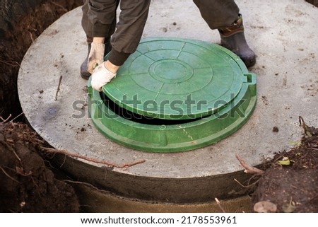 A worker installs a sewer manhole on a septic tank made of concrete rings. Construction of sewerage networks for country houses. Royalty-Free Stock Photo #2178553961