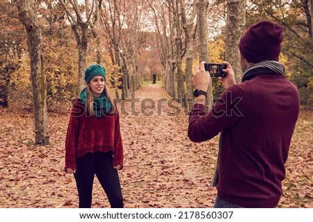 White Caucasian boy with his back turned taking a picture of his girlfriend with his smartphone on a ground of fallen brown leaves and a path of trees in a park in autumn